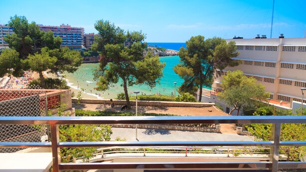 A coastal view from a balcony overlooking a beach with clear turquoise waters, a promenade lined with trees, and a hotel building in the background. - Spacious apartment with sea views and pool - UHC MARINA MAR (Salou)