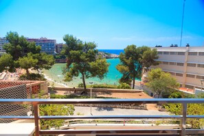 Une vue sur la côte, prise depuis un balcon donnant sur une plage aux eaux turquoise et limpides, une promenade bordée d’arbres et, au loin, un bâtiment d’hôtel.