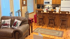 A cozy living room with a brown leather couch, wooden coffee table, and a laptop. The kitchen features wooden cabinets, a microwave, and a refrigerator. - Convenient Prime Location, Exceptional reviews!! We would love to host you. (Pigeon Forge)