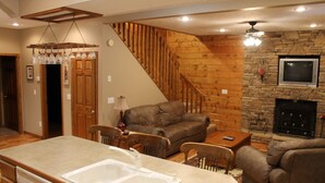 A cozy living area with a stone fireplace, wooden staircase, and a kitchen with a sink and dining table. - Convenient Prime Location, Exceptional reviews!! We would love to host you. (Pigeon Forge)