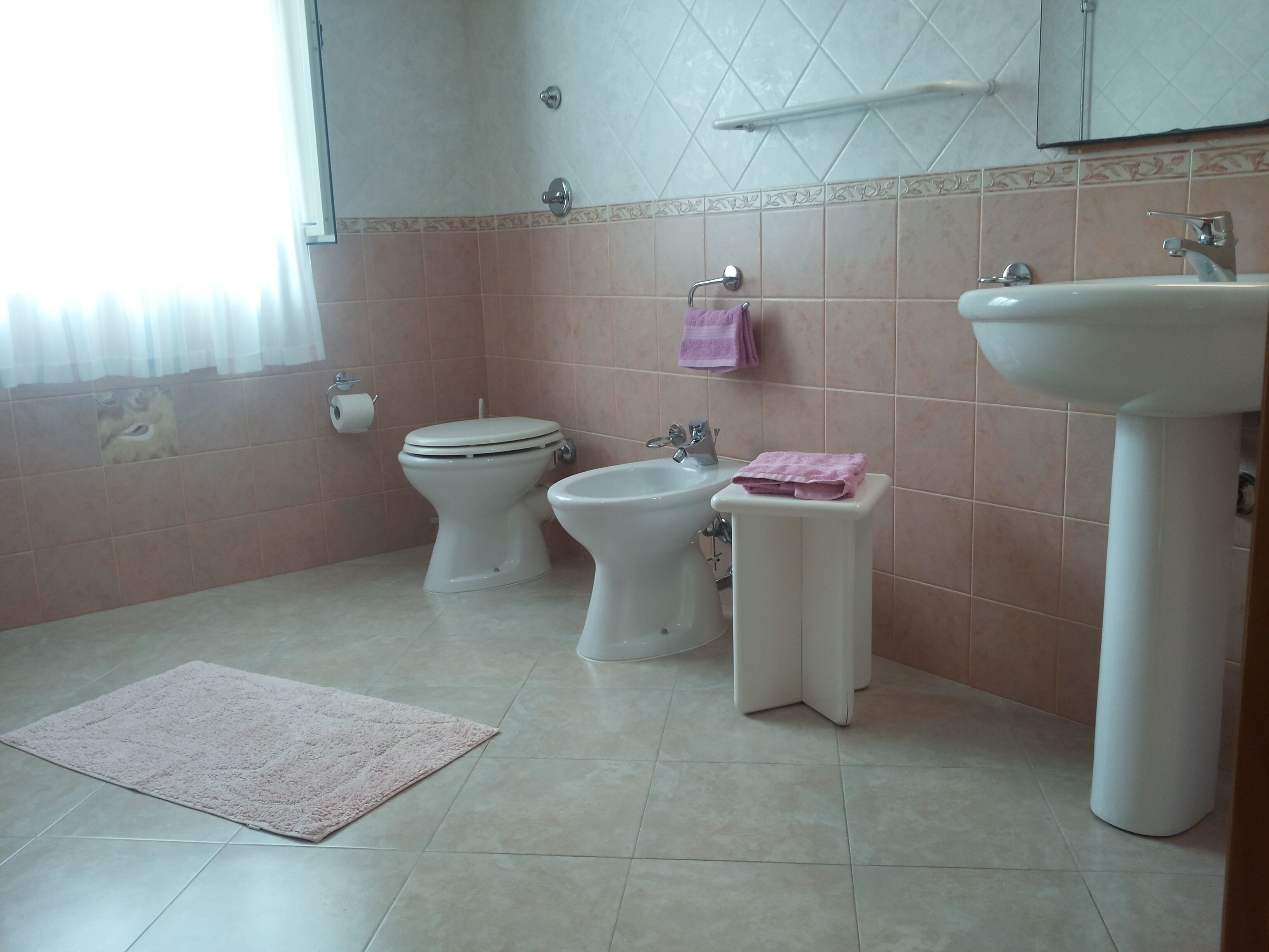 A bathroom with a toilet, bidet, sink, and a pink towel on a small stool.