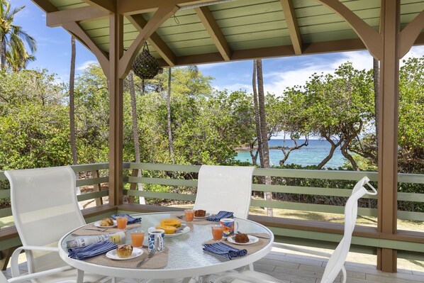 A covered outdoor seating area with a table set for breakfast, overlooking a tropical landscape with trees and a view of the sea.