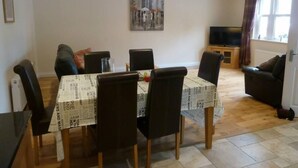 Ein Essbereich mit einem Tisch, bedeckt mit einem Tischlaufwerk mit Zeitungsdruck, umgeben von Stühlen. Im Hintergrund befindet sich eine Couch und ein Fernseher.