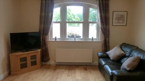Ein Wohnzimmer mit Fernseher auf einer Holzkommode, einem schwarzen Ledersofa und einem Fenster mit Vorhängen.
