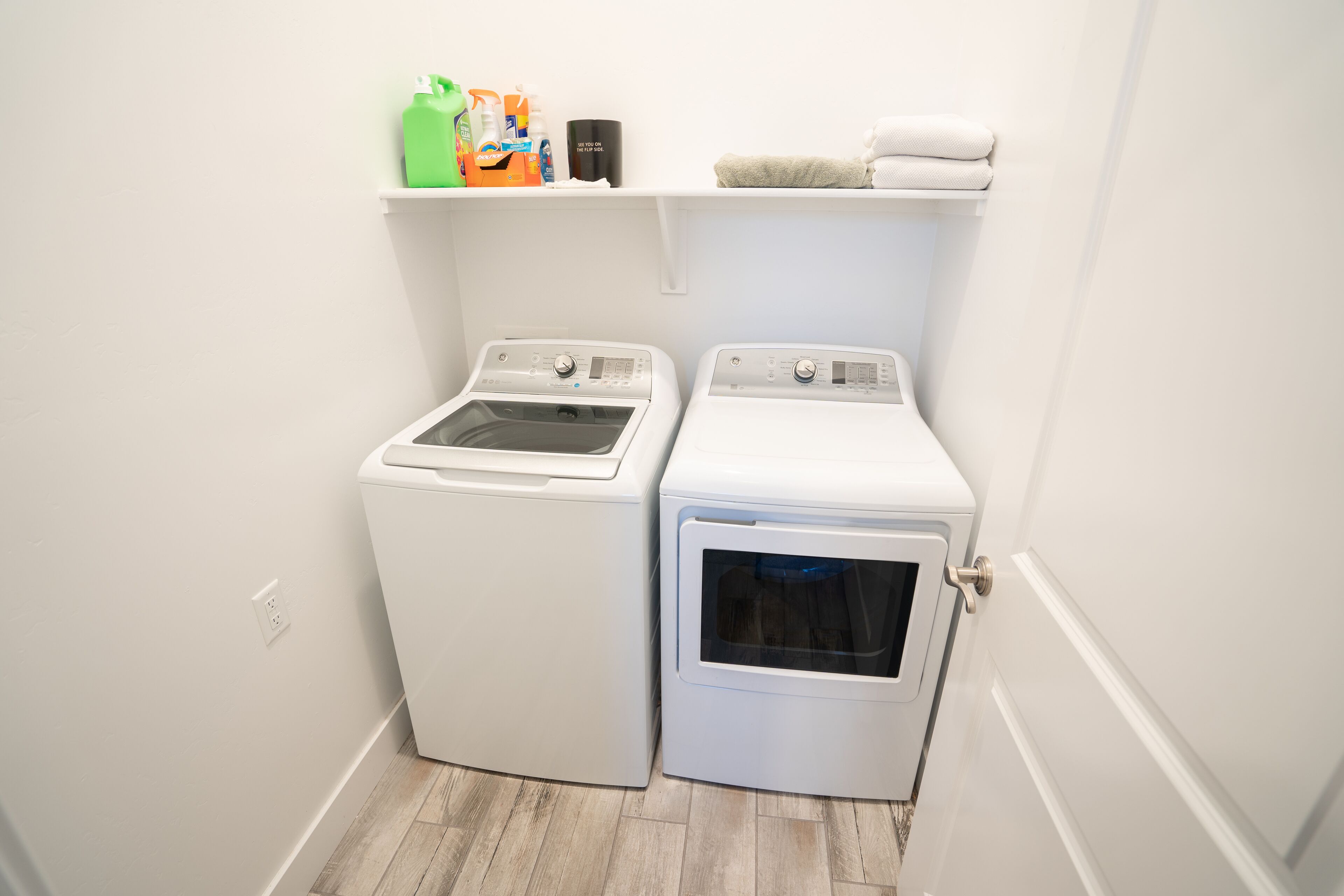 A laundry room with two washing machines and a shelf above containing cleaning supplies.