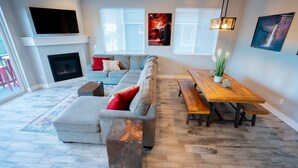 Een moderne woonkamer met een hoekbank, een open haard, een houten eettafel en een tv boven de open haard.