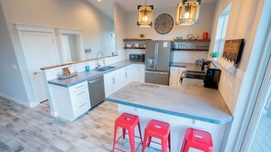 Een moderne keuken met witte kasten, een centraal eiland met rode krukken, een roestvrijstalen koelkast en hangende lampen.