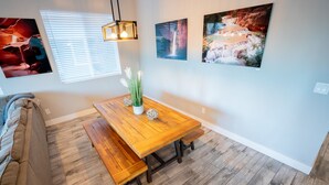 Een eetgedeelte met een houten tafel en bankjes, een sofa, en drie ingelijste foto’s aan de muur.