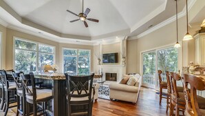 A spacious living room with a fireplace, a dining area with a large table and chairs, and a kitchen area with a view of the outdoors.