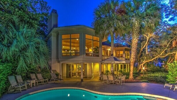 A two-story house with a swimming pool, surrounded by palm trees and outdoor furniture.