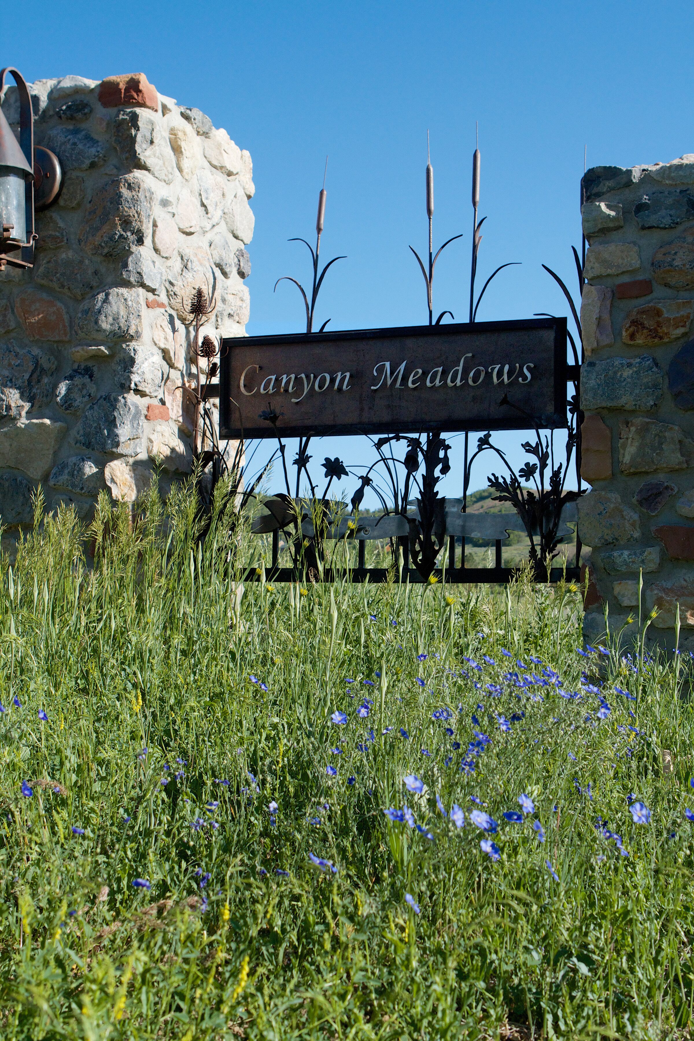 A rustic stone wall with a sign reading "Canyon Meadows" amidst a field of wildflowers.
