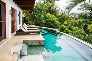 A swimming pool with a stone edge, surrounded by lush greenery and a white building with large windows and wooden shutters.