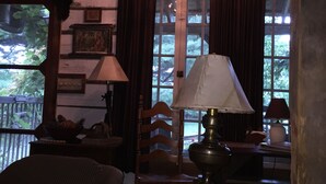 A cozy room with a rustic wooden ceiling, a large window with curtains, a wooden chair, a table with a lamp, and a decorative wheel light fixture.