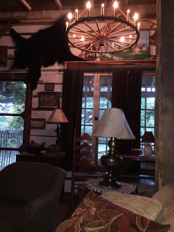 A cozy room with a rustic wooden ceiling, a large window with curtains, a wooden chair, a table with a lamp, and a decorative wheel light fixture. - Cabin rental near Cedar Point, Lake Erie, with Hot Tub, Campfire Pit, Gas Grill (Bellevue)