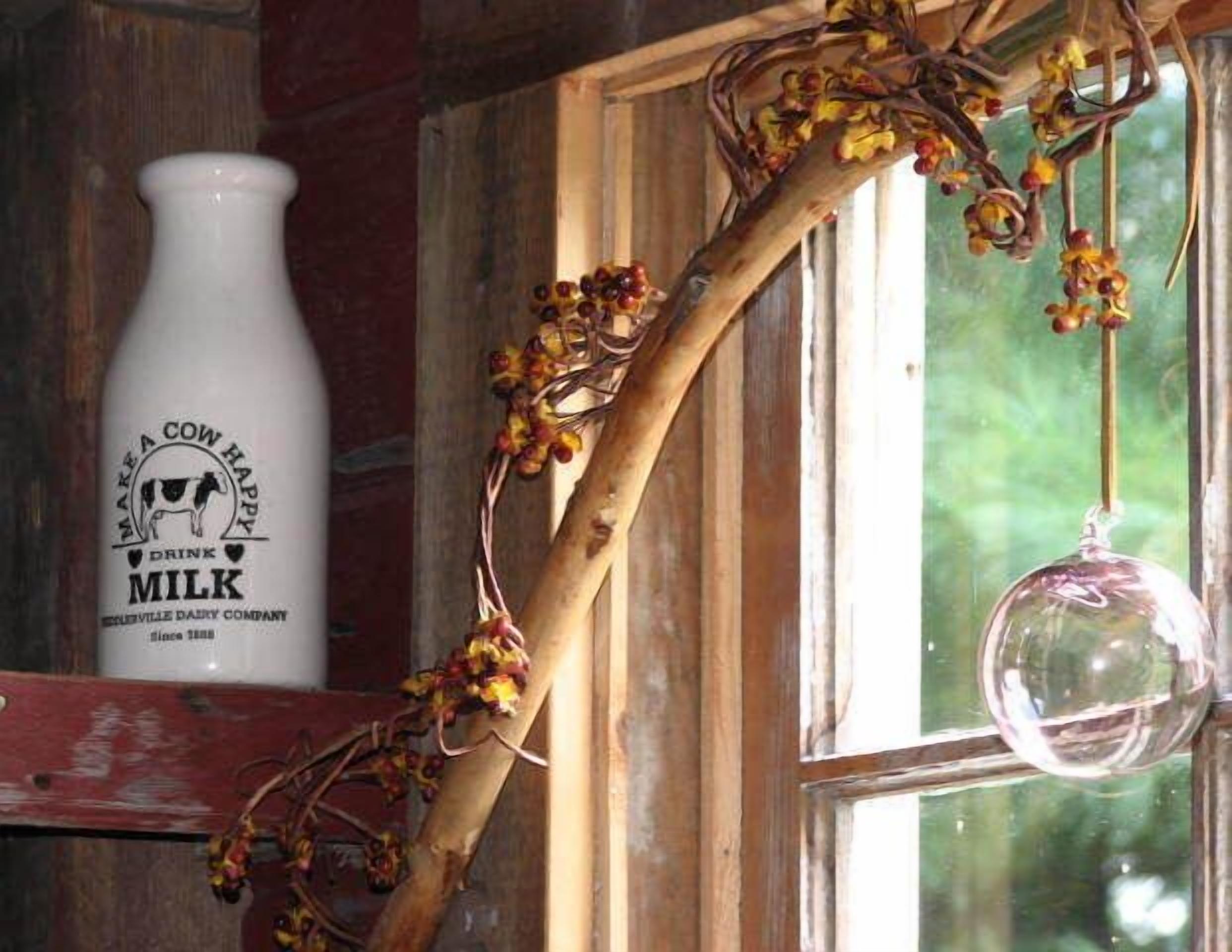 Un interior rústico con un marco de ventana de madera, una botella de leche blanca con la etiqueta 'MAKE A COW HAPPY MILK' y un adorno de vidrio colgante.