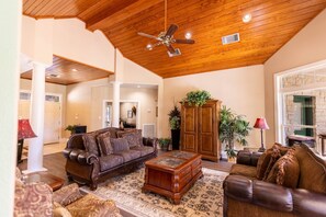 A spacious living room with a high ceiling, wooden beams, a large brown leather sofa, a wooden coffee table, and a potted plant. - Star Ranch Main Estate (Wimberley)
