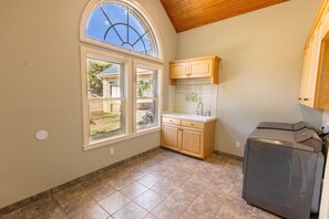 A kitchen with a large window, wooden cabinets, a washing machine, and a tiled floor. - Star Ranch Main Estate (Wimberley)
