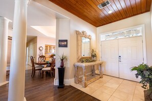 A hallway with a wooden ceiling, a white door, a decorative mirror, and a dining area with chairs and a table.