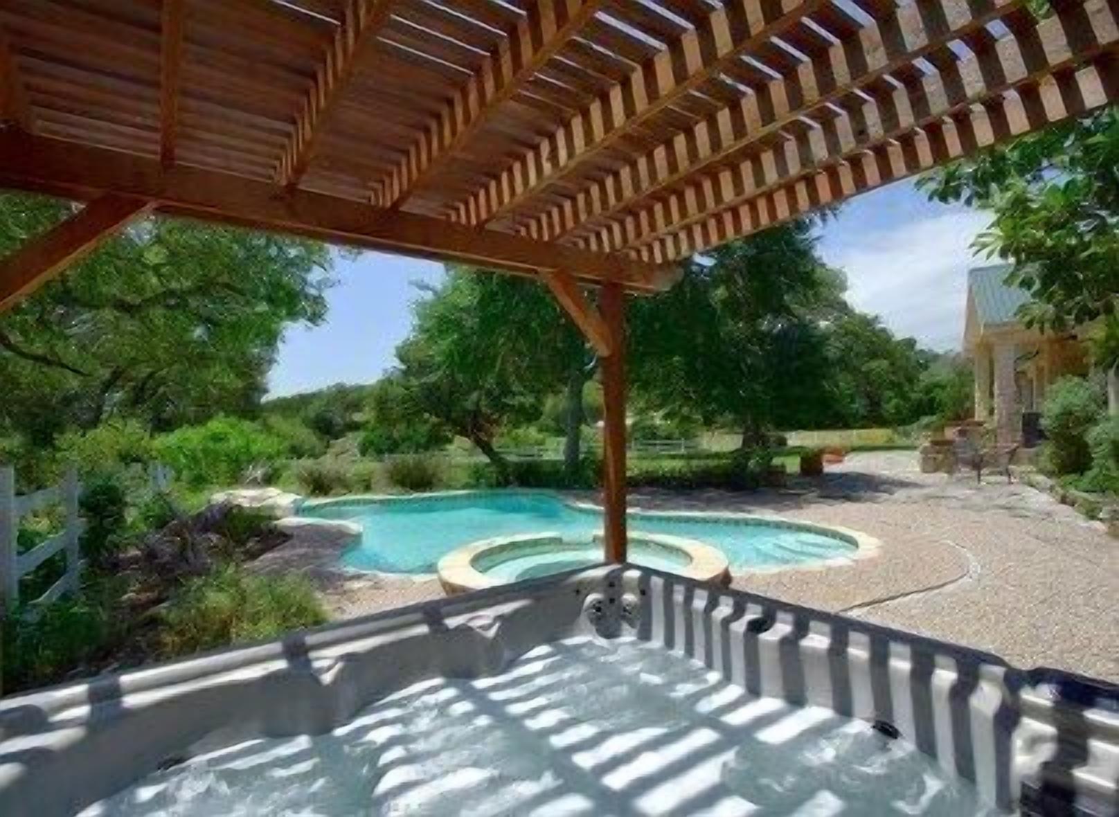 A hot tub under a wooden pergola with a pool in the background.