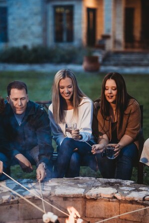 Three people sitting by a stone fire pit outdoors at dusk. - Star Ranch Main Estate (Wimberley)