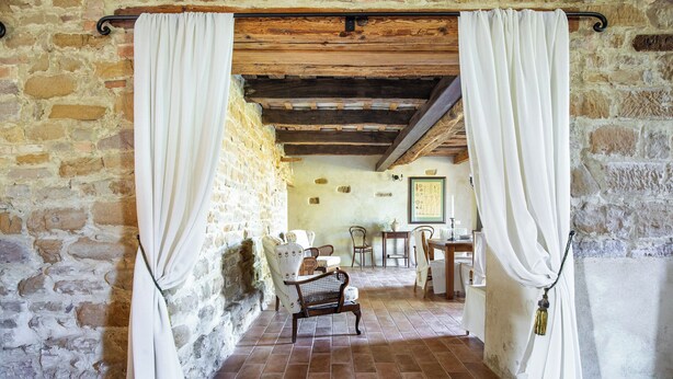 Una stanza rustica con pareti in pietra, soffitto in legno, una panca e un tavolo con sedie.