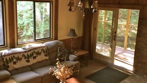 Ein gemütliches Wohnzimmer mit Holzbalkendecke, einer Couch mit gemustertem Überwurf, einem Couchtisch mit einer Vase und großen Fenstern mit Blick auf Grünflächen.