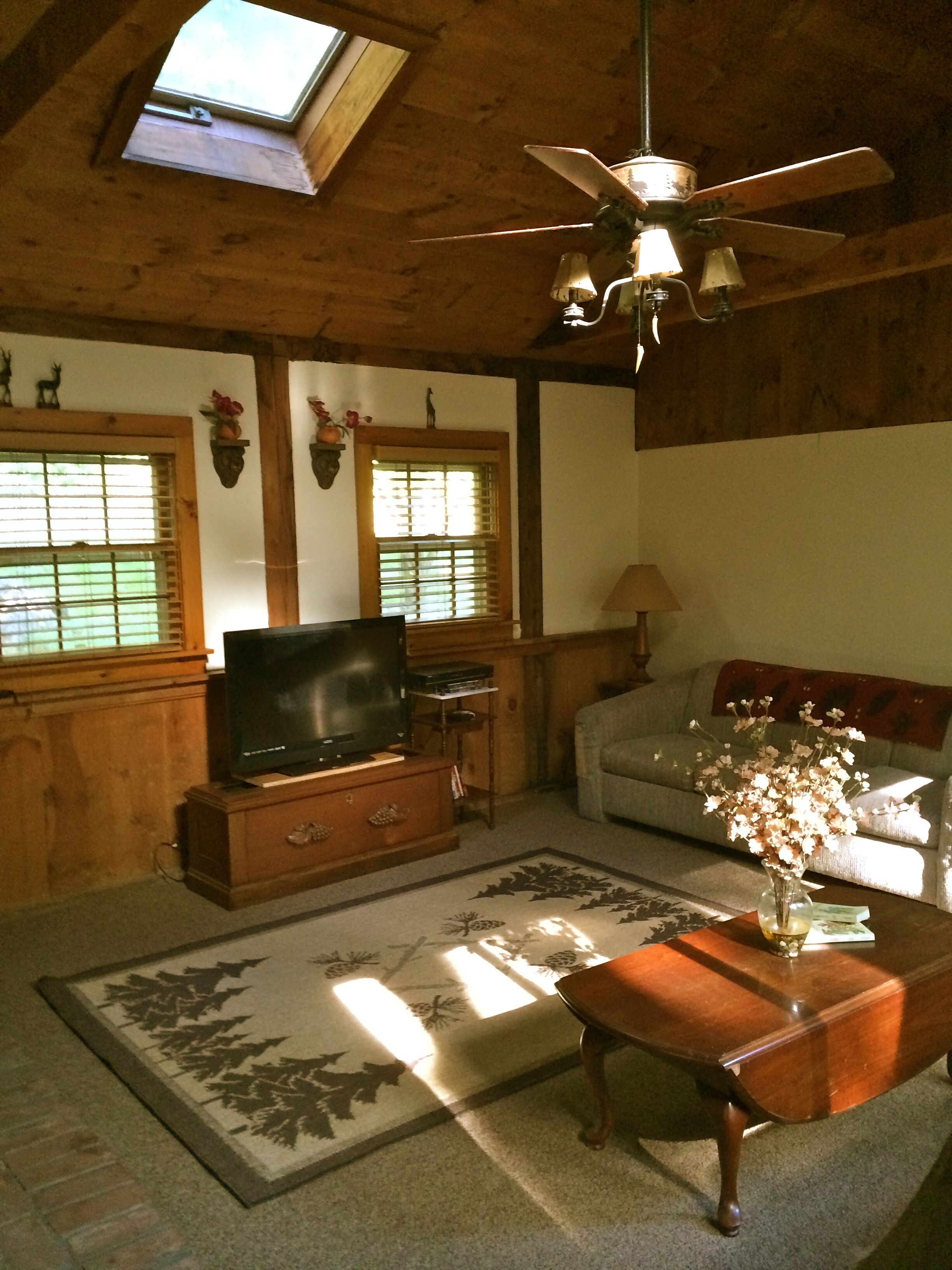 A cozy living room with a television, wooden furniture, a ceiling fan, and a window with blinds.