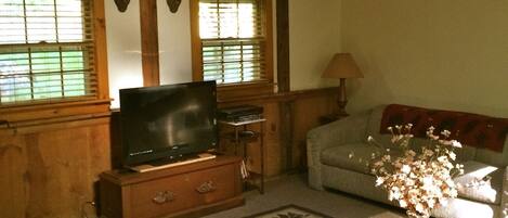 A cozy living room with a television, wooden furniture, a ceiling fan, and a window with blinds.