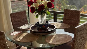 A glass-top dining table with wicker chairs, a vase with red and white flowers, and a view of a garden and mountains through large windows.