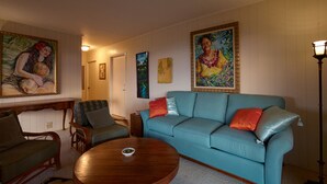 A living room with a teal sofa, a wooden coffee table, and a green armchair. There are paintings on the walls and a lamp on a side table.