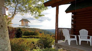 Une terrasse en bois avec deux chaises blanches donnant sur un paysage pittoresque : une colline sur laquelle se trouve un nichoir.