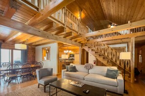 A spacious living area with a high ceiling, wooden beams, a sofa, a coffee table, and a dining area with chairs and a table. - The MOST private, beautiful Log Home for up to 12 guests. 10 min to Dreams Park (Hartwick)