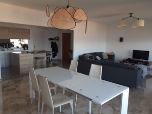Uma cozinha moderna com uma ilha central, uma área de jantar com mesa e cadeiras brancas, um sofá de cor escura e uma televisão de tela plana.