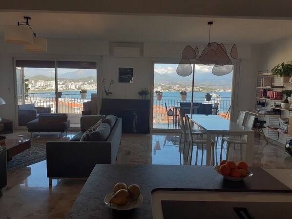 Ampla sala de estar com uma grande janela com vista para a cidade, área de jantar com mesa e cadeiras, e varanda com plantas em vasos.