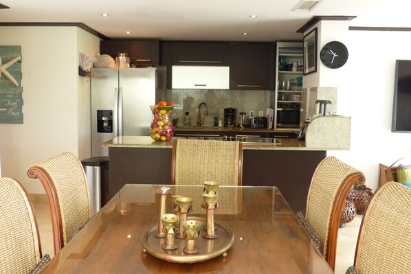 A modern kitchen with dark cabinetry, a stainless steel refrigerator, and a dining area with a glass table and wicker chairs. - SPACIOUS CORNER UNIT ON GROUND FLOOR (Paynes Bay)
