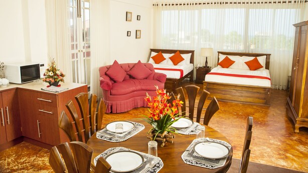A hotel room with a dining area, a bed with orange pillows, a wooden cabinet, and a ceiling fan. - Peaceful Harmonious Condo Equipped Kitchen Two Bath Three Bedrooms Aircondition (Mount Lavinia)