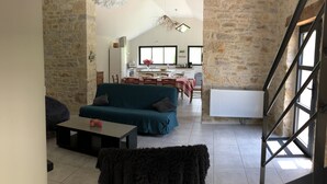 A cozy living room with a sofa, a coffee table, and a furry rug. A staircase leads to a kitchen area with a dining table and chairs.