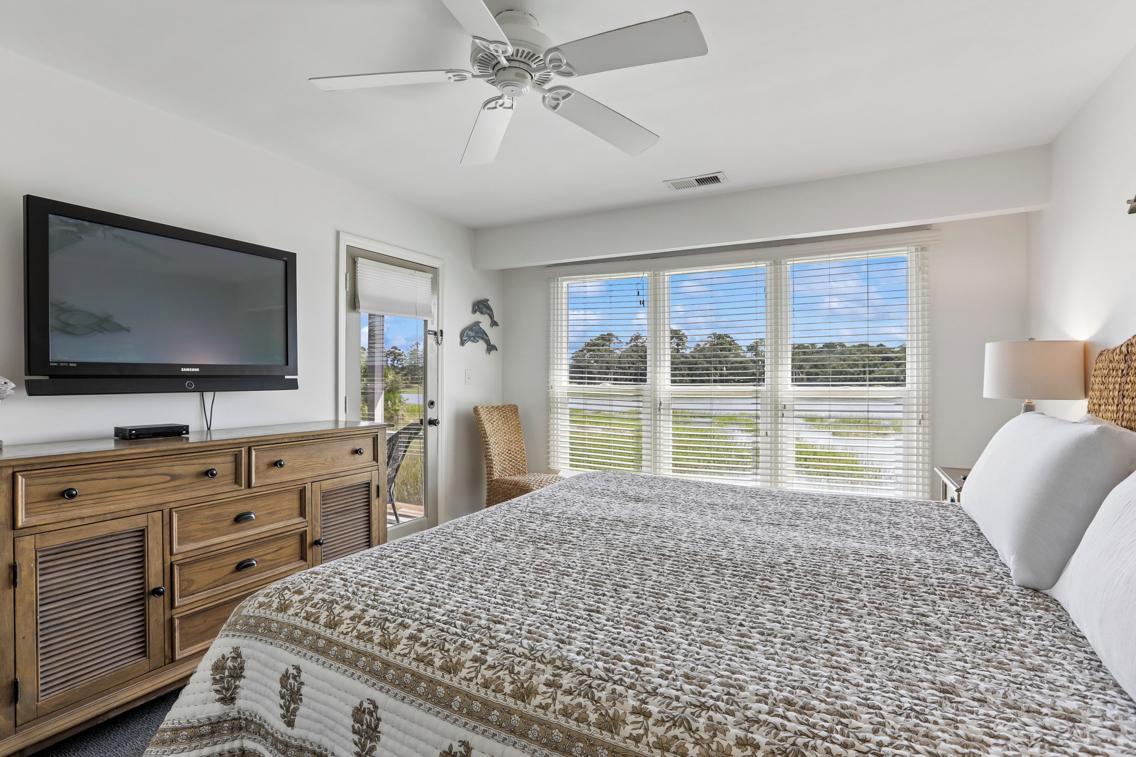 A bedroom with a bed, a television, a ceiling fan, a window with blinds, and a view of the outdoors.
