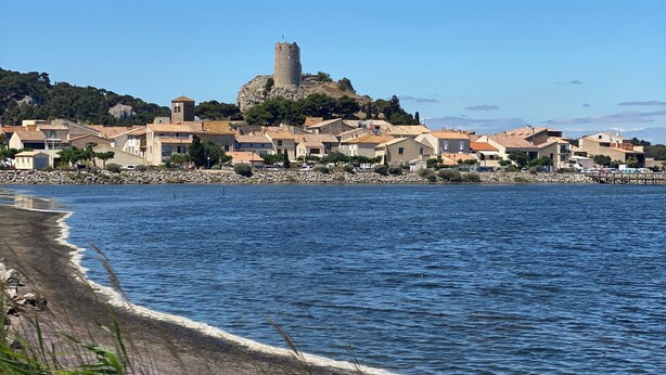 Uma vila costeira com uma torre imponente numa colina, uma praia com ondas e edifícios com telhados de terracota.