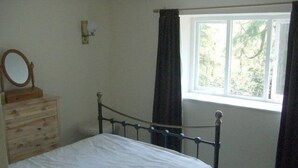 Ein Schlafzimmer mit einem hölzernen Schminktisch, einem Bett mit Metallgestell, einem Fenster mit Vorhängen und einem an der Wand befestigten Spiegel.