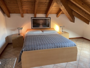 Une chambre avec un plafond en bois, un lit recouvert d’une couverture matelassée, deux lampes de chevet, une table de chevet, une commode et un tableau encadré accroché au mur.