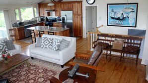 A spacious living area with a white sofa, a coffee table, and a wooden bench. A kitchen with wooden cabinets and stainless steel appliances is visible in the background. - Beautiful Oceanfront Home in Estate Setting Gloucester — FALL RENTAL AVAILABLE! (Gloucester)