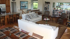 A spacious living room with a white sofa, a coffee table, and a dining area with a wooden table and chairs. There is a kitchen area with a refrigerator and a clock on the wall. - Beautiful Oceanfront Home in Estate Setting Gloucester — FALL RENTAL AVAILABLE! (Gloucester)