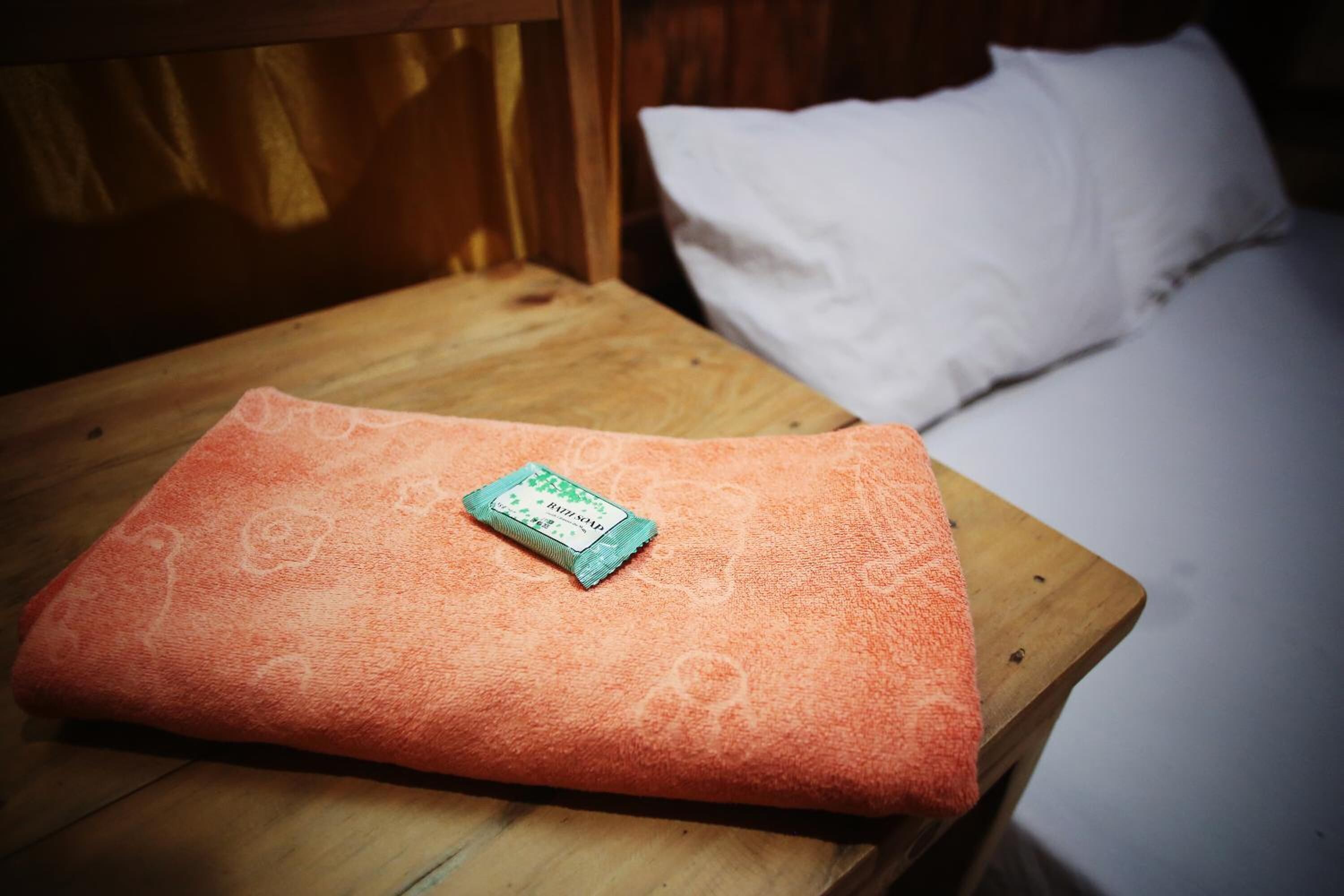 A neatly folded orange towel with a packet on a wooden table beside a bed with white pillows.