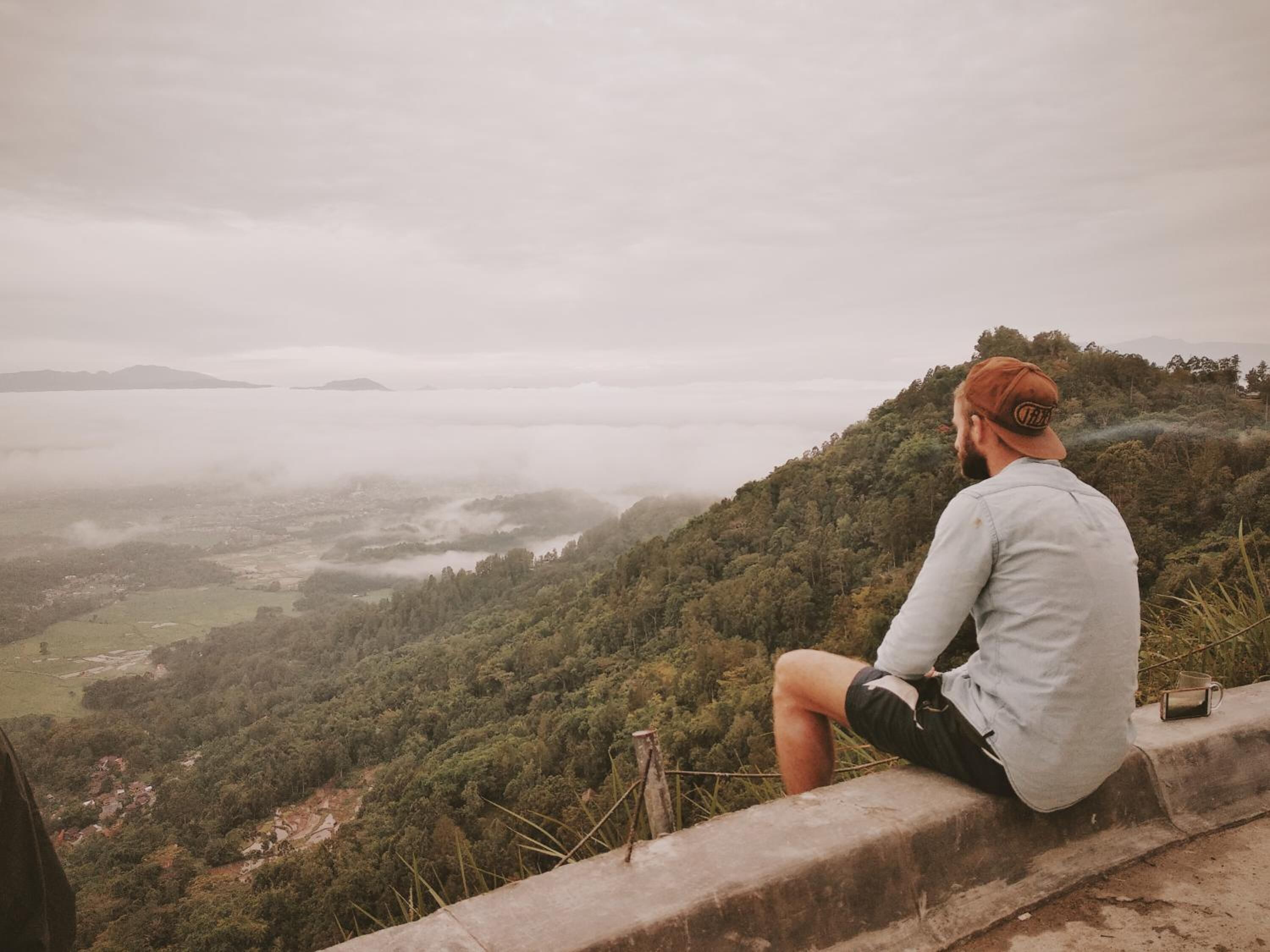 A person sitting on a concrete ledge overlooking a valley with dense greenery and a misty atmosphere.