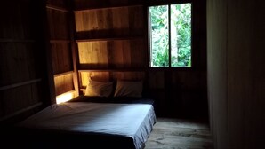 Une petite pièce à la lumière tamisée, avec un lit simple, des murs en bois et une fenêtre donnant sur la verdure.