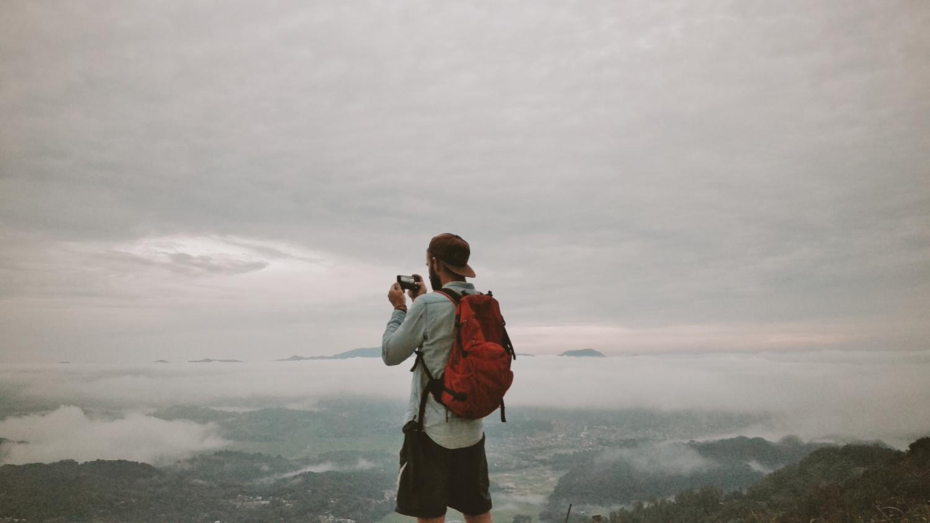Une personne prend une photo avec un appareil photo, porte un sac à dos rouge et se tient sur une colline surplombant une vallée.