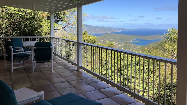 A covered outdoor area with two chairs and a table, overlooking a scenic landscape with hills and a body of water. - Sea Cottage - Charming and Romantic with a Stunning View (St. John)