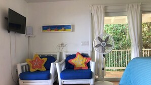 A room with two white armchairs with blue cushions and star-shaped pillows, a wooden coffee table with a book, a TV mounted on the wall, and a ceiling fan. - Sea Cottage - Charming and Romantic with a Stunning View (St. John)