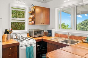 A compact kitchen with a gas stove, a built-in oven, a toaster, and a coffee maker. There are wooden cabinets and a window with a view of greenery. - Sea Cottage - Charming and Romantic with a Stunning View (St. John)